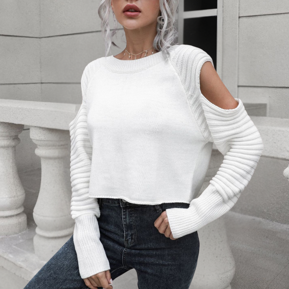 Vrouw in witte trui met open schouders en geribbelde mouwen, gecombineerd met donkere jeans, poseert voor een stenen balustrade.