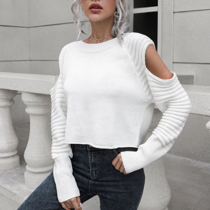 Vrouw in witte trui met open schouders en geribbelde mouwen, gecombineerd met donkere jeans, poseert voor een stenen balustrade.
