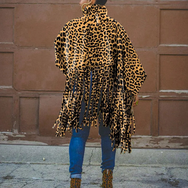 Vrouw draagt een luipaardprint poncho met franjes, gecombineerd met blauwe jeans en luipaardlaarzen, tegen een bruine achtergrond. Modieuze herfstkleding.