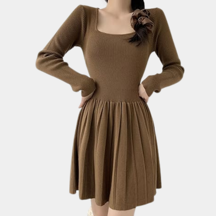 Bruine gebreide jurk met lange mouwen en geplooide rok, versierd met bloemdetail op de schouder. Perfect voor herfstmode en casual elegantie.