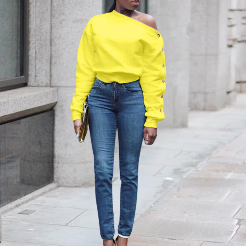 Vrouw in gele off-shoulder trui en blauwe skinny jeans, staand op trottoir. Modieuze casual outfit, stedelijke achtergrond.