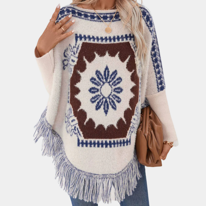 Vrouw draagt een gebreide poncho met blauw en bruin geometrisch patroon en franjes, gecombineerd met een bruine tas. Perfect voor herfstmode.