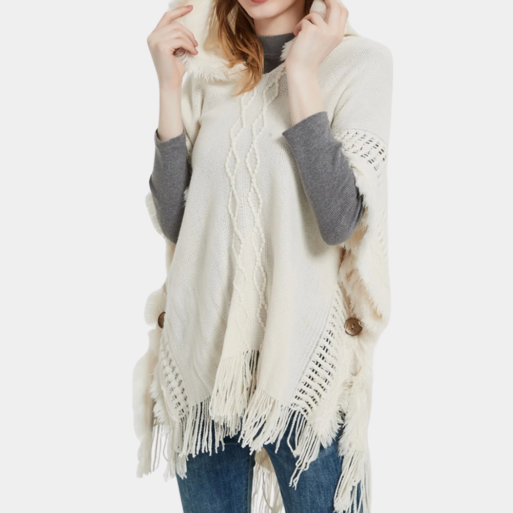 Vrouw draagt een crèmekleurige gebreide poncho met franjes en kabelpatroon, gecombineerd met een grijze trui en blauwe jeans. Modieuze herfstkleding.