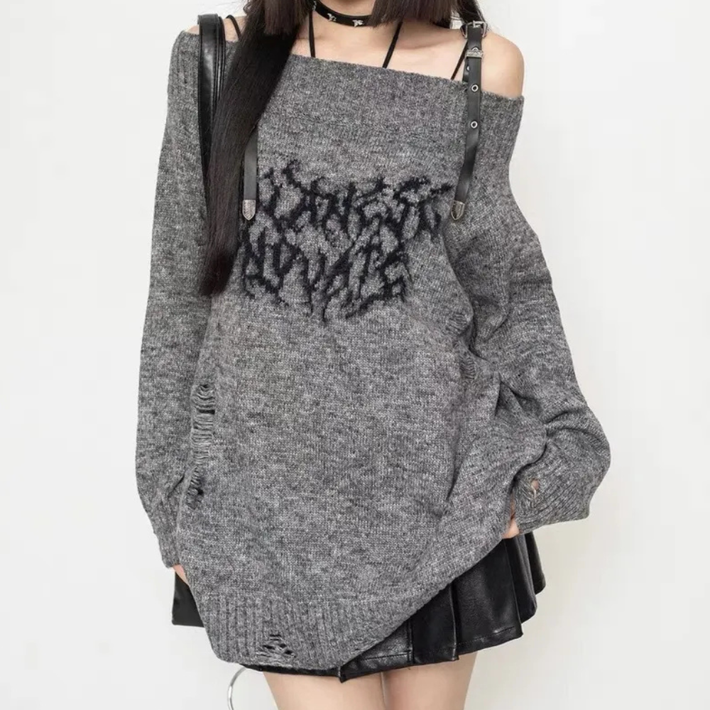 Grijze oversized trui met zwarte grafische print, off-shoulder ontwerp, leren riemen en gerafelde details, gedragen door model. Trendy streetwear.