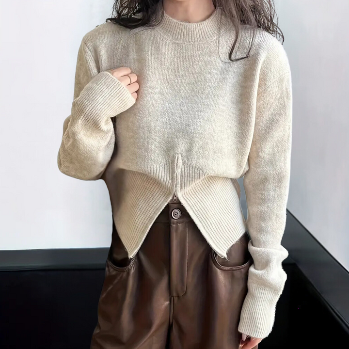 Vrouw draagt beige trui met unieke asymmetrische zoom, gecombineerd met bruine leren broek. Modieuze dameskleding, herfst-winter stijl.