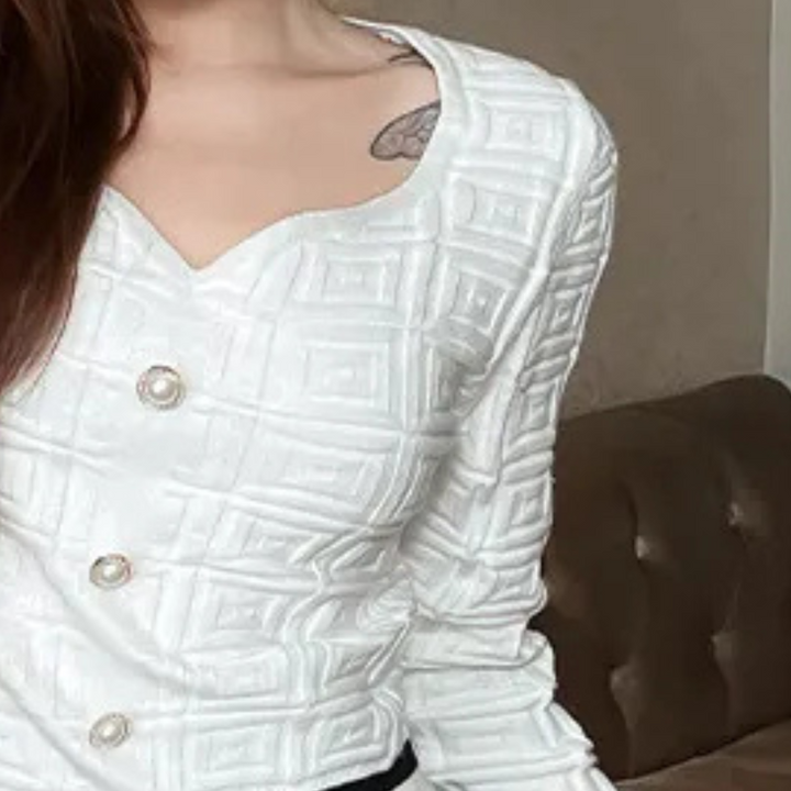 Vrouw in witte, gewatteerde blouse met vierkante patronen en knopen, zittend op een bruine bank. Stijlvolle damesmode, elegant design.