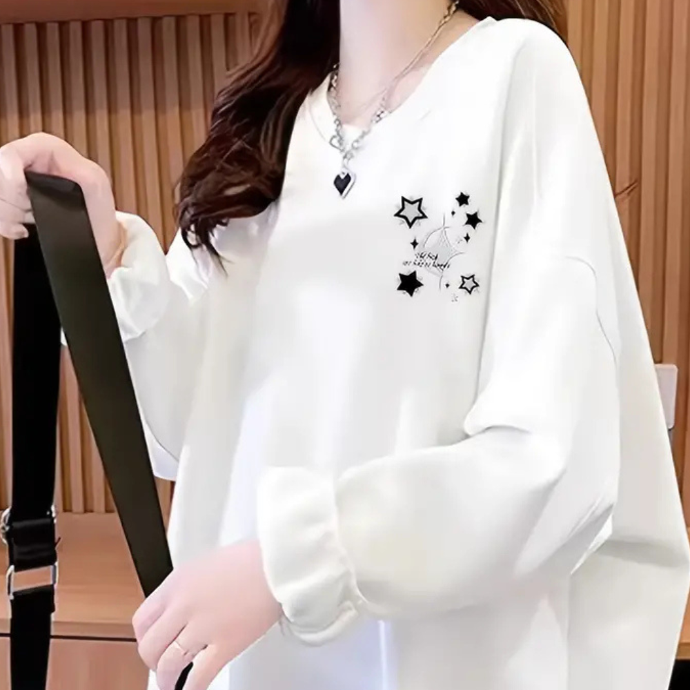 Vrouw in witte oversized trui met sterrenprint en hartketting, houdt zwarte schoudertas vast. Modieuze dameskleding, casual stijl.