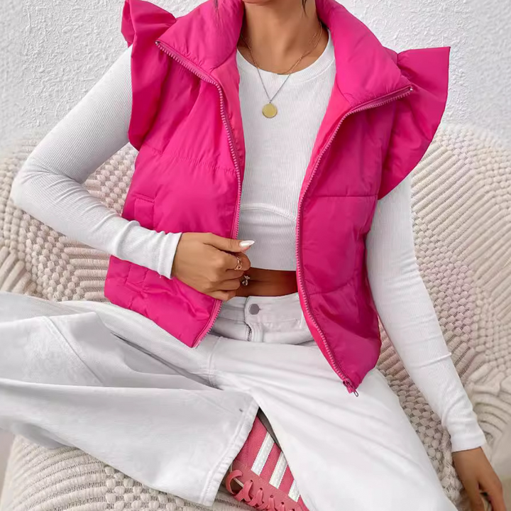 Vrouw in roze gewatteerd vest met ruches, witte trui en broek, zittend op een beige bank. Modieuze dameskleding, casual outfit, trendy stijl.