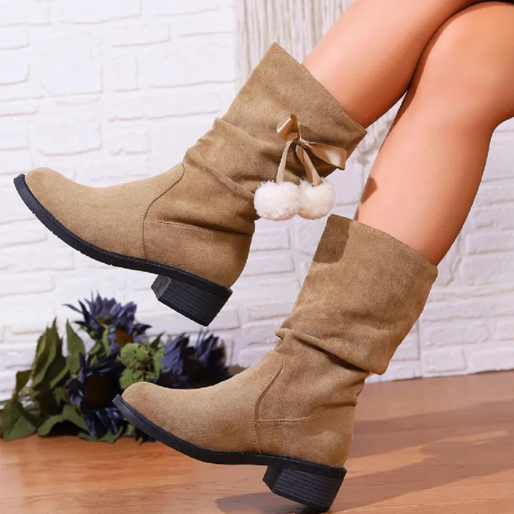 Beige suède laarzen met pompons, lage hak, tegen witte bakstenen muur. Trendy damesmode, comfortabele herfst- en winterlaarzen.