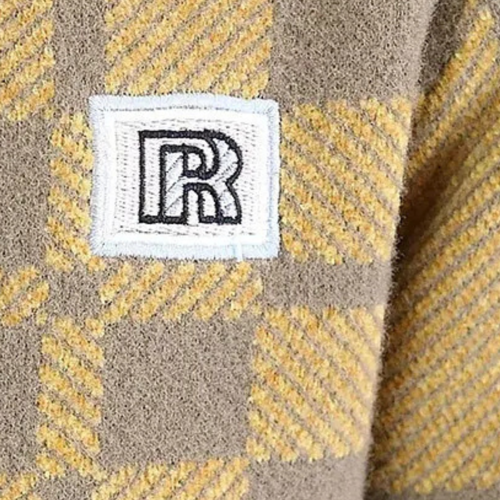 Geruit beige en geel wollen stof met geborduurd wit en zwart "R" logo, close-up. Modieuze textiel, herfstkleding, luxe design.