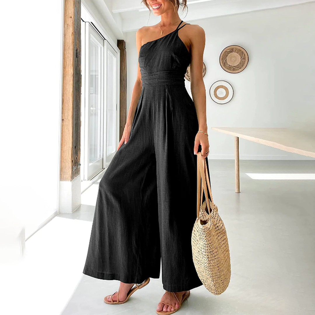 Vrouw in zwarte jumpsuit met halternek, draagt een strooien tas, staat in moderne, lichte kamer. Zomeroutfit, casual mode, dameskleding.