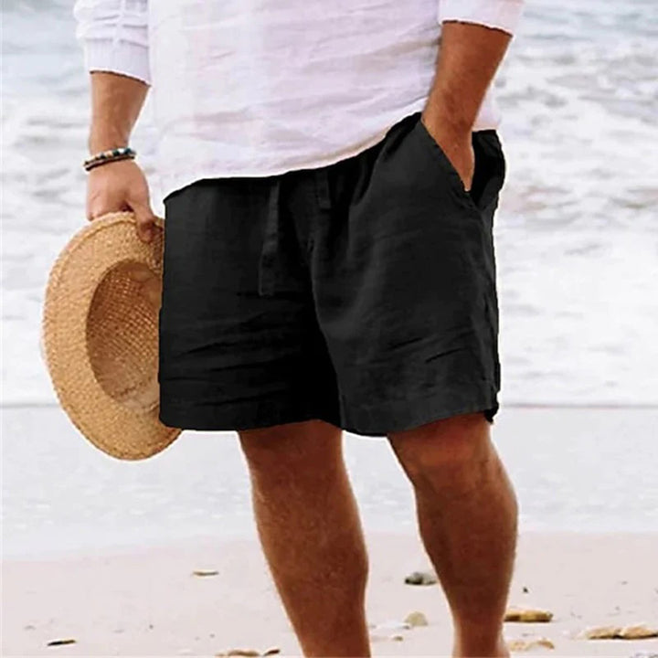 Man in zwarte linnen korte broek en wit overhemd, houdt strohoed vast op strand. Zomerse herenkleding, casual strandoutfit, comfortabele stijl.