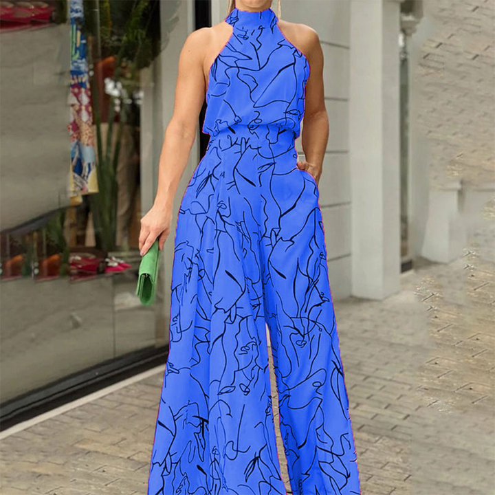 Vrouw in blauwe jumpsuit met abstracte print, halternek en wijde pijpen, poseert buiten. Trendy damesmode, zomeroutfit, stijlvolle kleding.