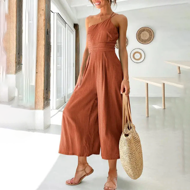 Vrouw in bruine, asymmetrische jumpsuit met strooien tas in moderne, lichte kamer. Zomermode, casual outfit, trendy kledingstijl.
