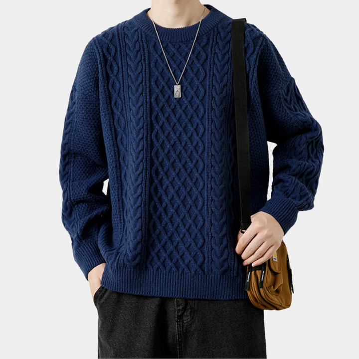 Man in blauwe kabeltrui met ketting en bruine tas. Modieuze herenkleding, casual winteroutfit, comfortabele gebreide trui.