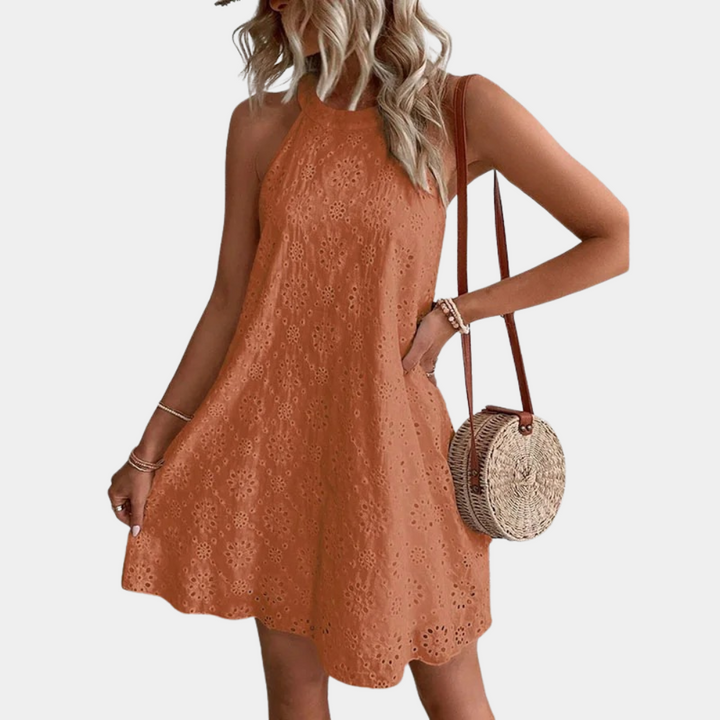 Oranje zomerjurk met bloemenpatroon, mouwloos en halternek. Vrouw draagt een ronde rieten tas. Perfect voor casual zomermode.