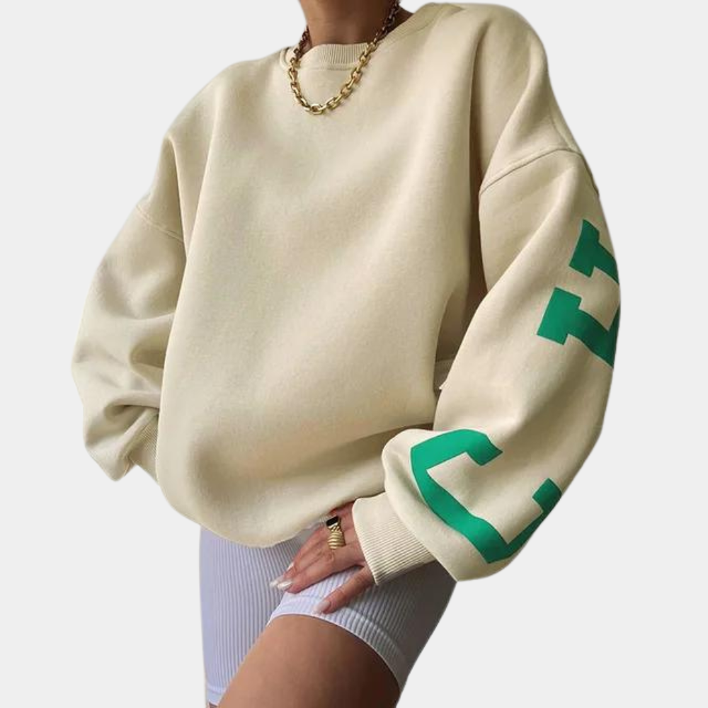 Beige oversized sweatshirt met groene letters op de mouw, gedragen door model met gouden ketting en witte shorts. Trendy streetwear mode.