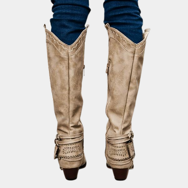 Beige leren dameslaarzen met hak, sierstiksels en gespen, gedragen over blauwe jeans. Stijlvolle kniehoge laarzen voor herfstmode.
