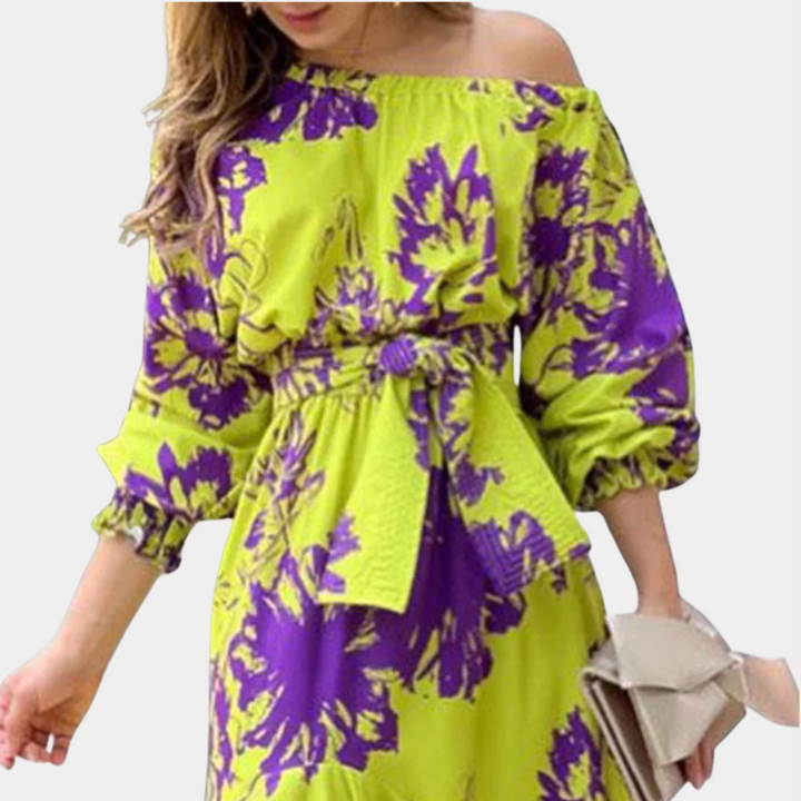 Vrouw in gele off-shoulder jurk met paarse bloemenprint, tailleband en lange mouwen, houdt beige clutch vast. Trendy zomeroutfit.