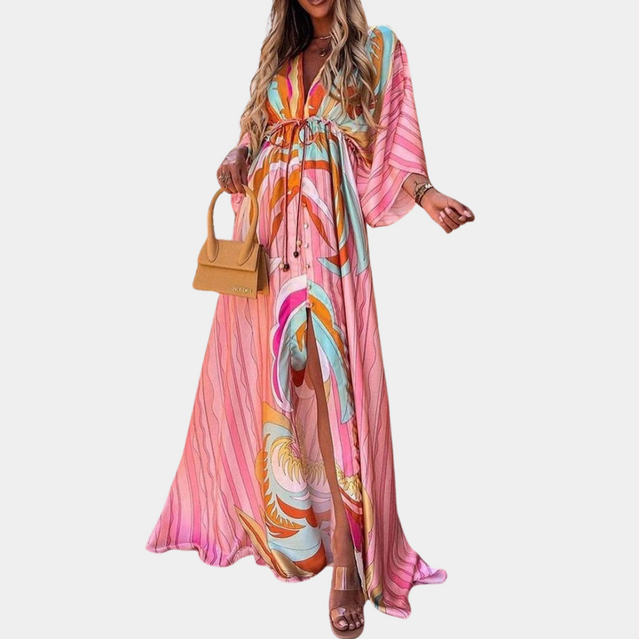 Vrouw in kleurrijke roze maxi-jurk met abstract patroon, draagt een beige handtas en sandalen. Perfect voor zomermode en boho-stijl.