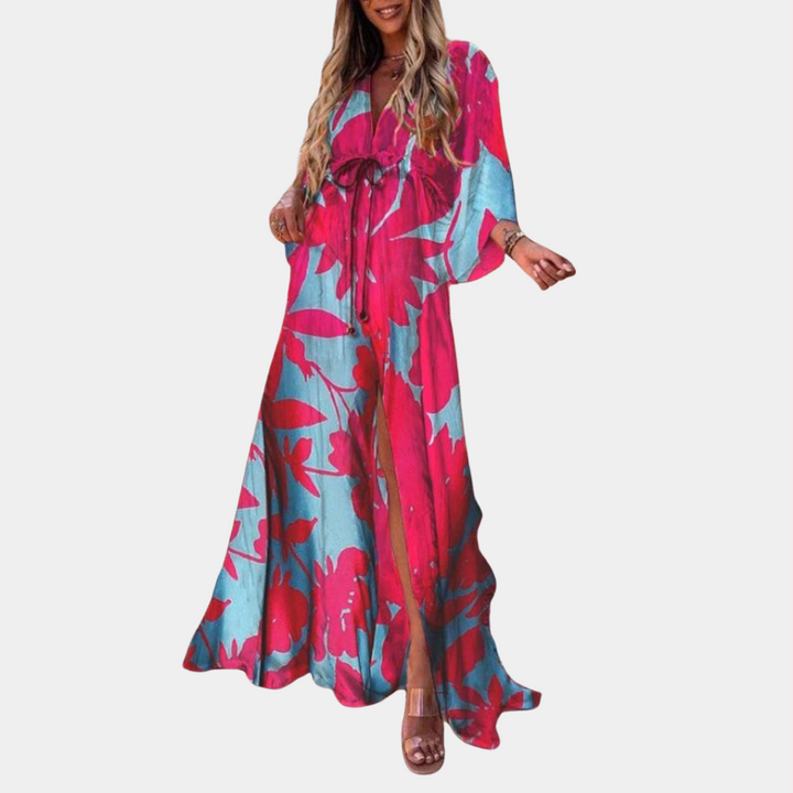Vrouw in lange, kleurrijke jurk met rood-blauw bloemenpatroon, wijde mouwen en striksluiting. Perfect voor zomermode en strandkleding.