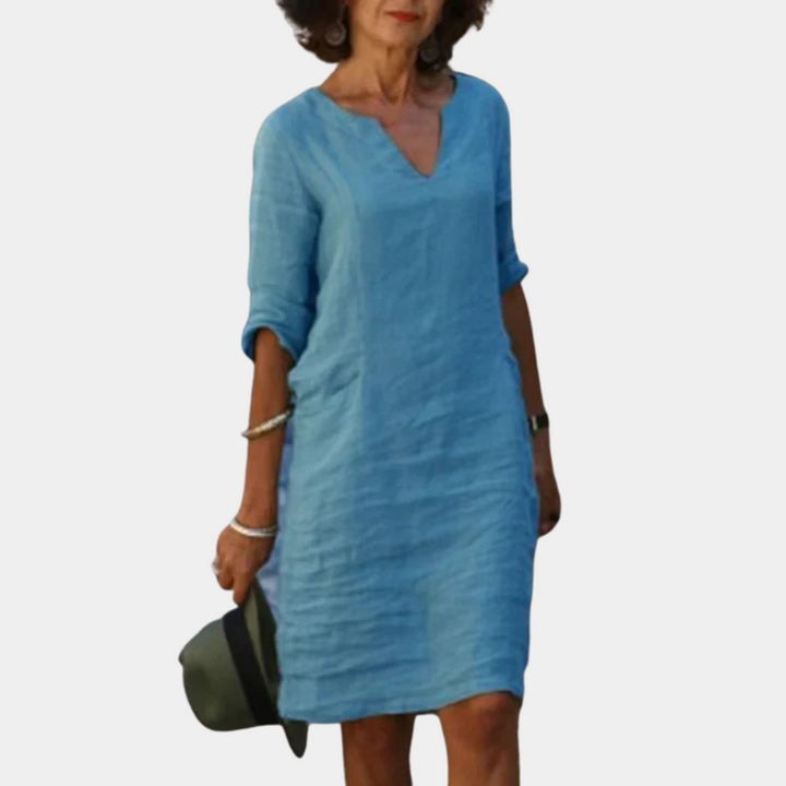 Vrouw in lichtblauwe linnen jurk met V-hals en opgerolde mouwen, houdt een hoed vast. Zomerse mode, casual stijl, comfortabele kleding.