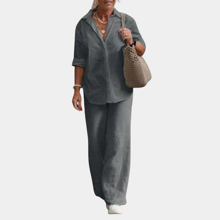 Vrouw in grijs linnen pak met lange mouwen, wijde broek en witte sneakers, draagt een beige gehaakte tas. Stijlvolle casual damesmode.