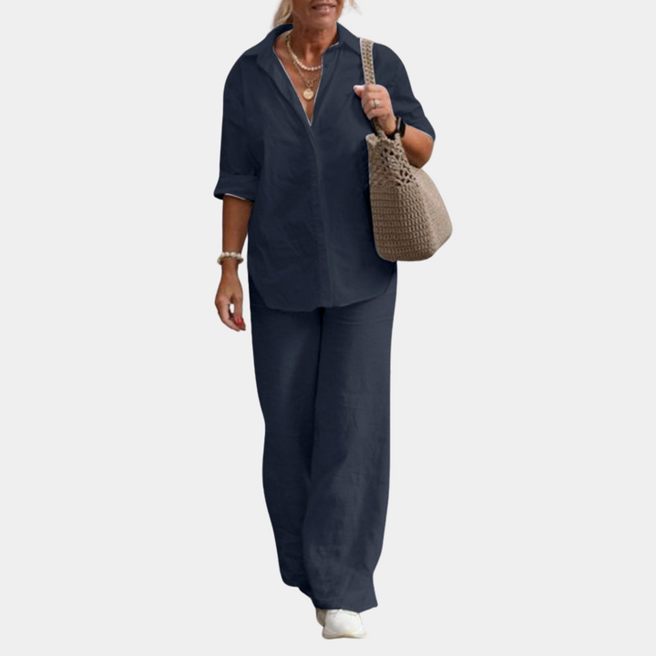 Vrouw in marineblauw linnen pak met opgerolde mouwen, draagt een beige gehaakte tas en witte sneakers. Stijlvolle damesmode, casual chic outfit.