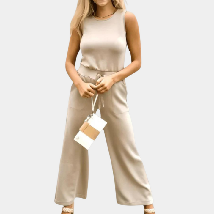 Vrouw in beige mouwloze jumpsuit met wijde pijpen, draagt een kleine witte en bruine schoudertas. Modieuze dameskleding, casual chic stijl.