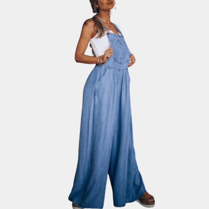 Vrouw in blauwe denim jumpsuit met wijde pijpen, witte tanktop en sandalen. Modieuze dameskleding, casual stijl, comfortabele pasvorm.