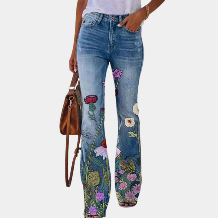 Vrouw draagt blauwe jeans met kleurrijke bloemenprint, gecombineerd met een witte top en bruine leren tas. Trendy damesmode, casual stijl.