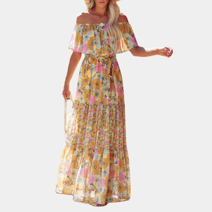 Vrouw in gele bloemenjurk met off-shoulder ontwerp, zomerse maxi-stijl, kleurrijk patroon, casual en elegant, perfect voor strand of vakantie.