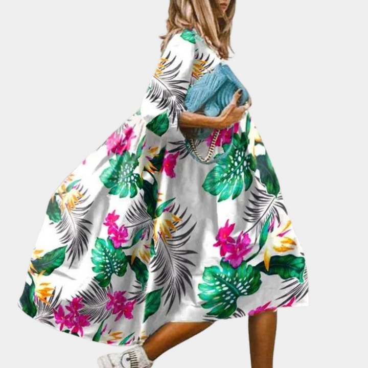 Vrouw in kleurrijke zomerjurk met tropische bloemenprint, draagt blauwe clutch en witte sneakers. Trendy damesmode, casual zomeroutfit.