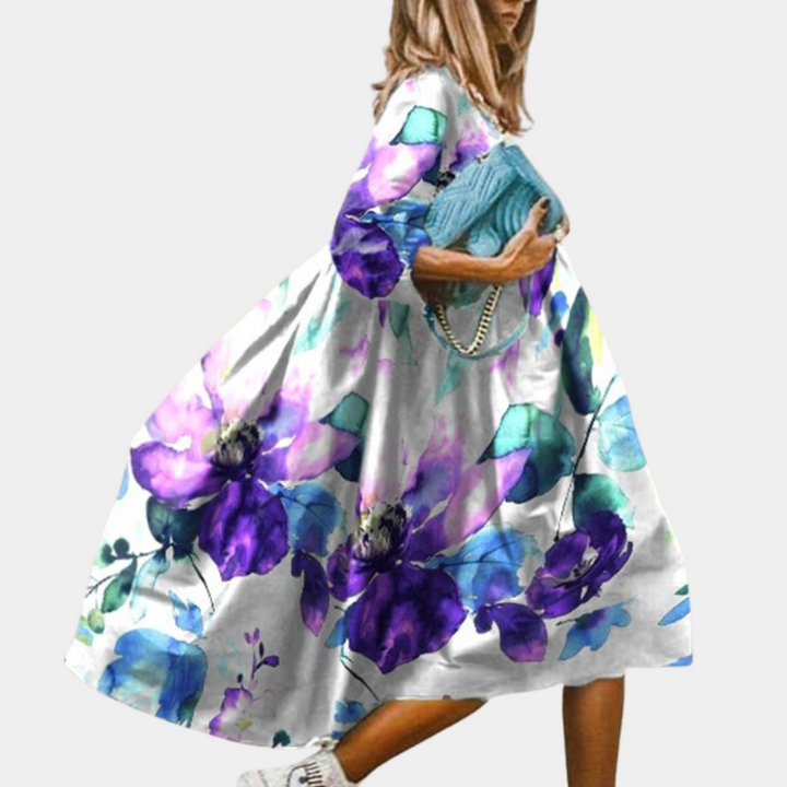 Vrouw in kleurrijke bloemenjurk met paarse en blauwe accenten, draagt een blauwe handtas. Trendy mode, casual stijl, lente/zomer outfit.