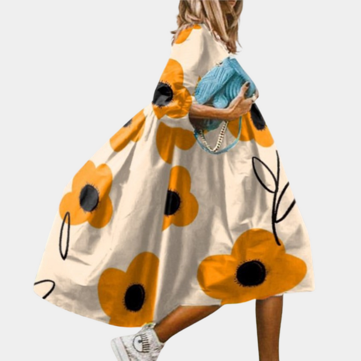 Vrouw in lange, gele bloemenjurk met grote oranje bloemen, draagt blauwe tas. Trendy zomerjurk, casual mode, dameskleding.