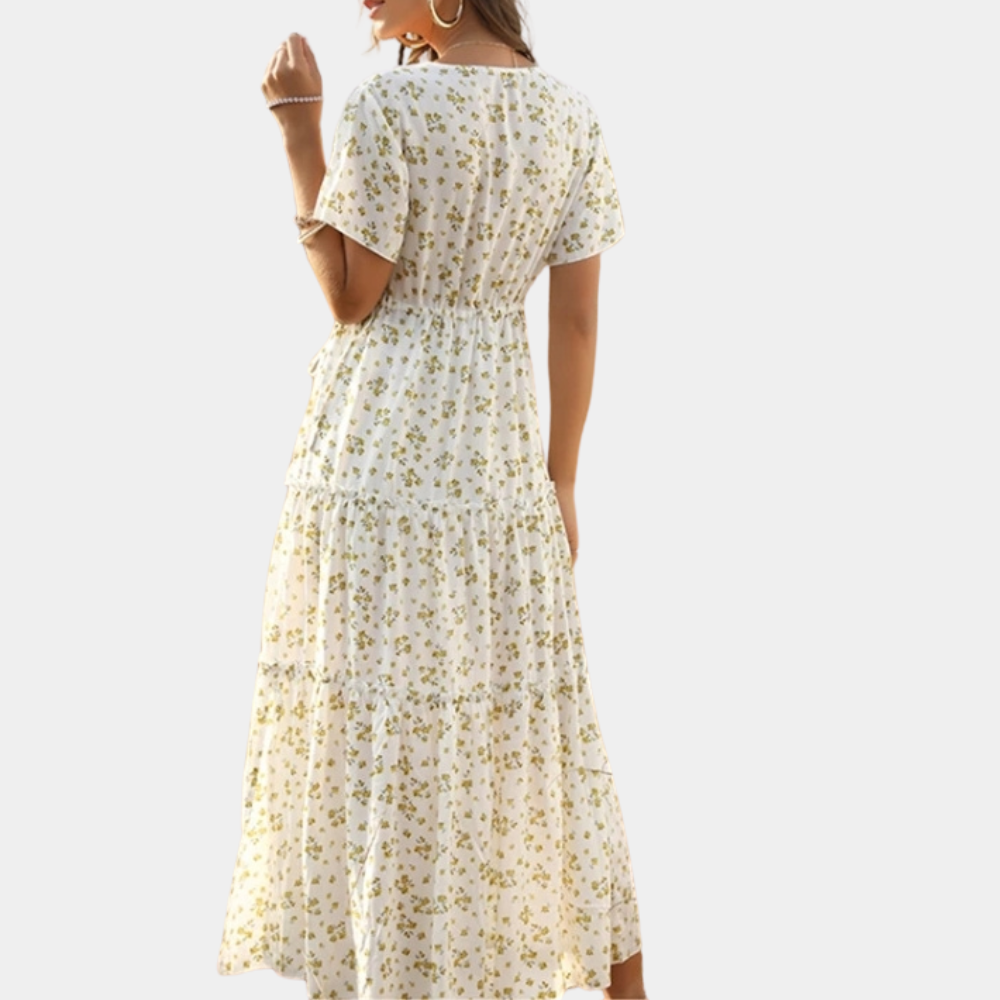 Vrouw in witte bloemenjurk met korte mouwen en tailleband, staand tegen een grijze achtergrond. Zomerse damesmode, casual elegantie.