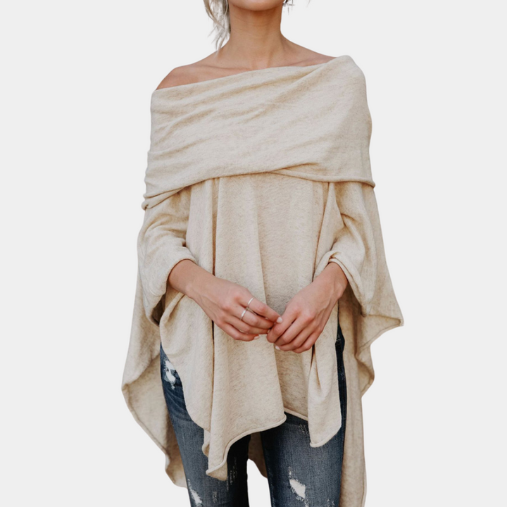 Vrouw draagt een off-shoulder, beige poncho trui met losse pasvorm, gecombineerd met gescheurde jeans. Modieuze dameskleding, casual stijl.