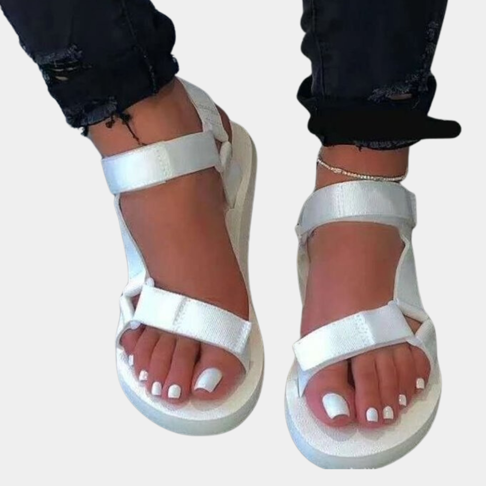 Witte sandalen met dikke zool en klittenbandsluiting, gedragen door een persoon met witte nagellak. Trendy zomerschoenen voor dames.