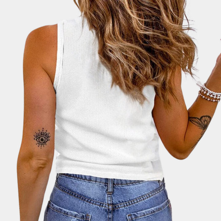 Vrouw in witte tanktop en blauwe jeans, met golvend bruin haar en armband, toont rug en tatoeage van oog op arm. Casual mode, zomeroutfit.
