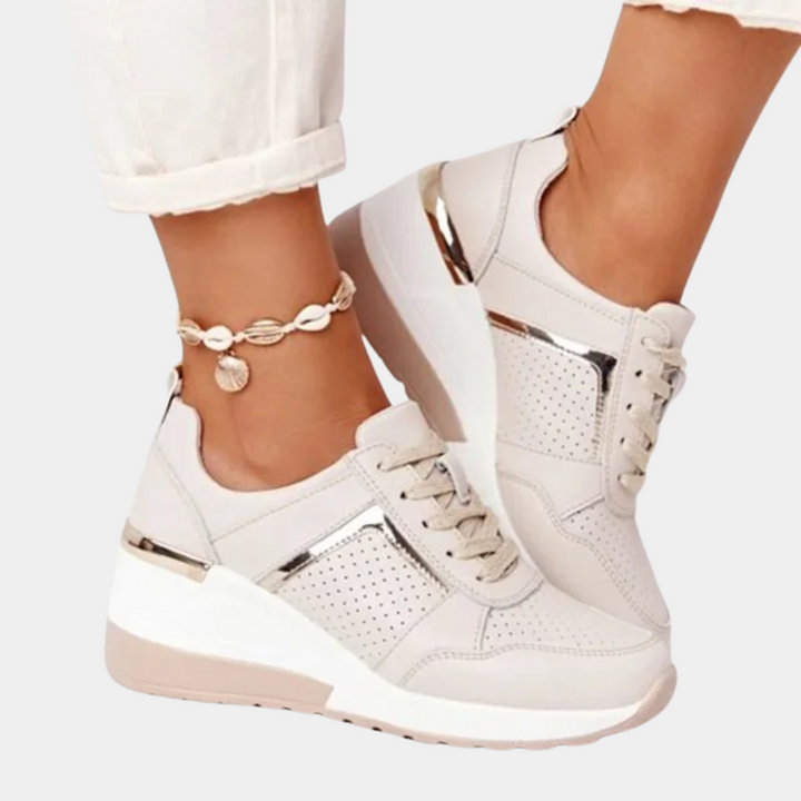 Beige sneakers met sleehak, metallic details en perforaties, gedragen met witte broek en schelpenenkelbandje. Trendy damesmode, comfortabele schoenen.