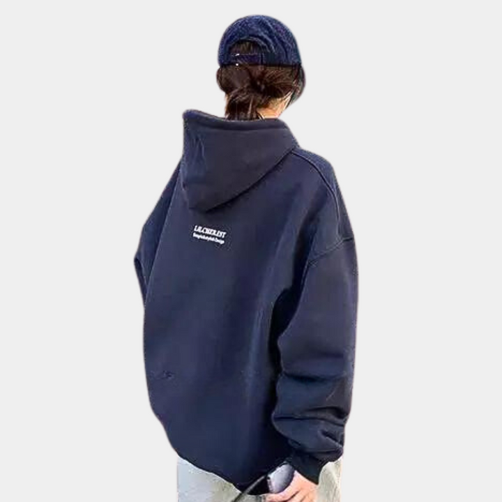 Persoon in oversized zwarte hoodie met capuchon en tekst "LACKLUST" op de rug, draagt een blauwe pet. Casual streetwear mode.
