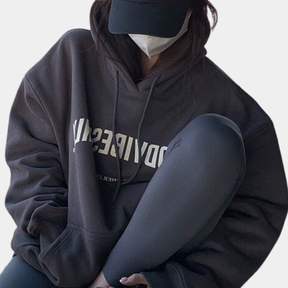 Donkergrijze oversized hoodie met capuchon, gedragen door persoon met zwarte pet en mondkapje. Comfortabele streetwear, casual mode.