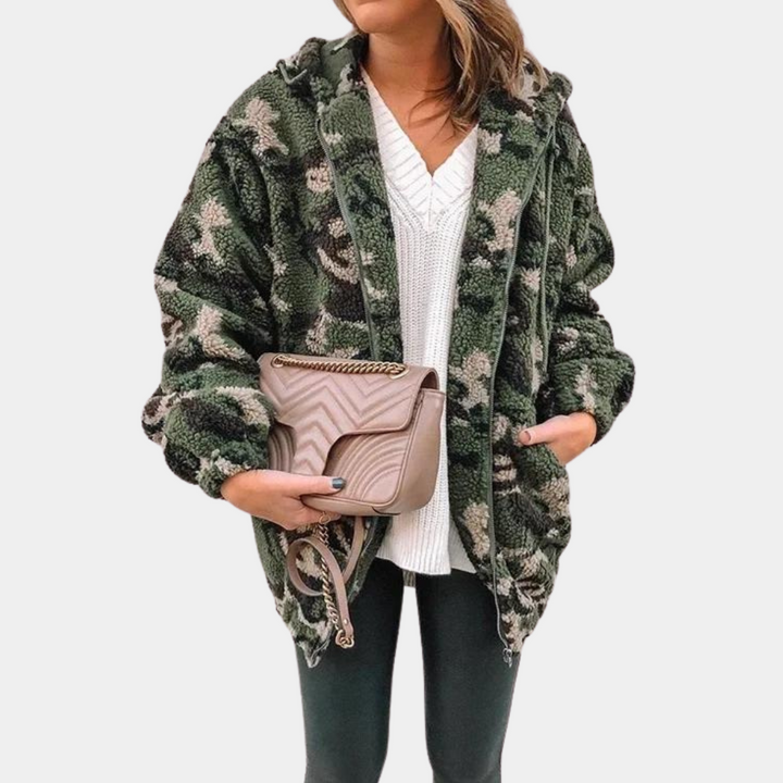 Vrouw in groene camouflage teddyjas met capuchon, witte trui en zwarte broek, houdt beige schoudertas vast. Modieuze winteroutfit, casual stijl.