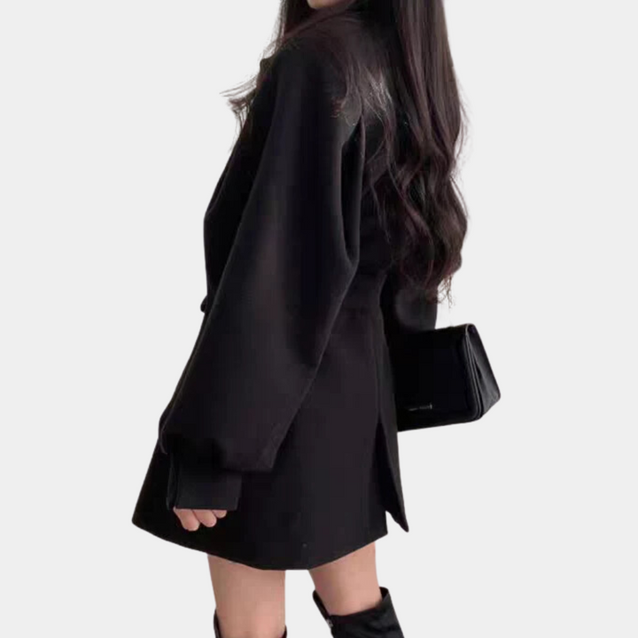 Zwarte damesjas met lange mouwen, getailleerd design, gedragen met zwarte handtas en laarzen. Modieuze herfst- of winterkleding.