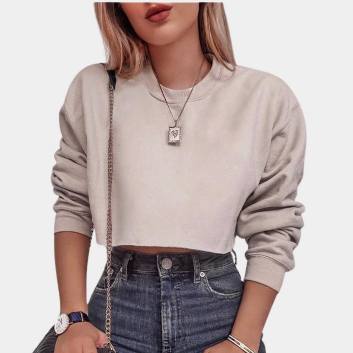 Vrouw in beige cropped sweatshirt, hoge taille jeans, ketting en horloge. Modieuze dameskleding, casual outfit, trendy streetwear.