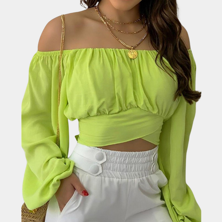Vrouw in limegroene off-shoulder blouse met lange mouwen en gouden kettingen, gecombineerd met witte broek. Trendy damesmode, zomeroutfit.