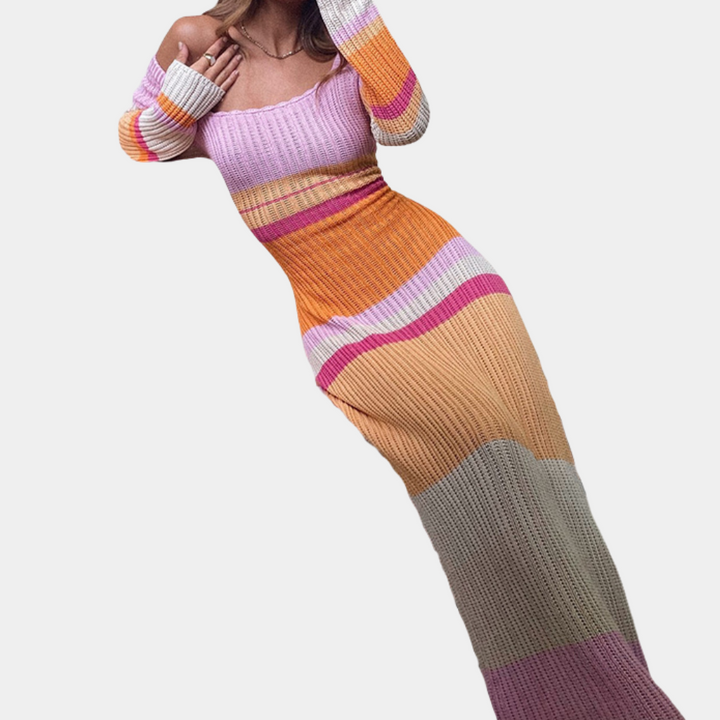 Vrouw in kleurrijke, gebreide off-shoulder maxi-jurk met strepen in roze, oranje en beige. Trendy damesmode, herfstkleding, stijlvolle outfit.