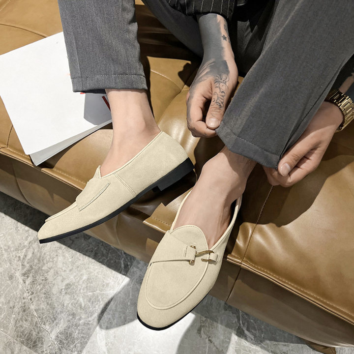 Beige heren loafers met strikdetail, gedragen door man in grijze broek op bruine bank. Stijlvolle casual schoenen voor moderne herenmode.