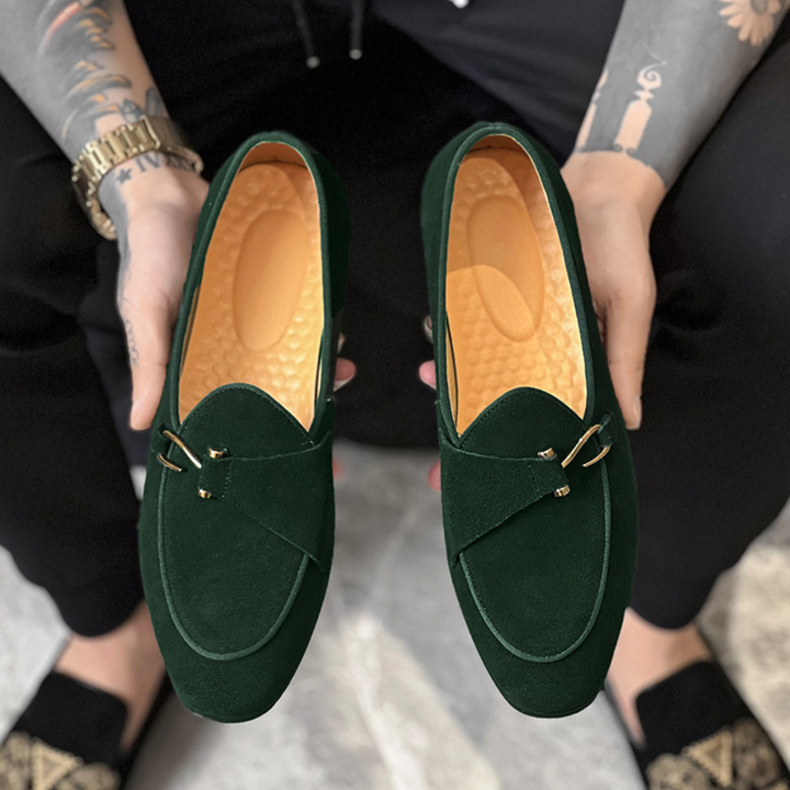 Groene suède loafers met gouden details, vastgehouden door getatoeëerde handen. Stijlvolle herenschoenen, perfect voor casual en formele gelegenheden.