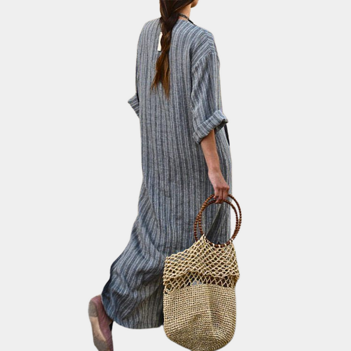 Grijze gestreepte maxi-jurk met lange mouwen, gedragen door vrouw met gevlochten rieten tas. Modieuze dameskleding, casual zomeroutfit.